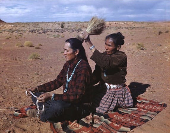 Navajo Barber Shop. An Indian women dresses her husband's hair as they sit on the desert floor in the open-air-living room. The man holds the string with which the women will bind his locks. Doing each others hair is a sign of affection for each other. Photo and Caption by Josef Muench